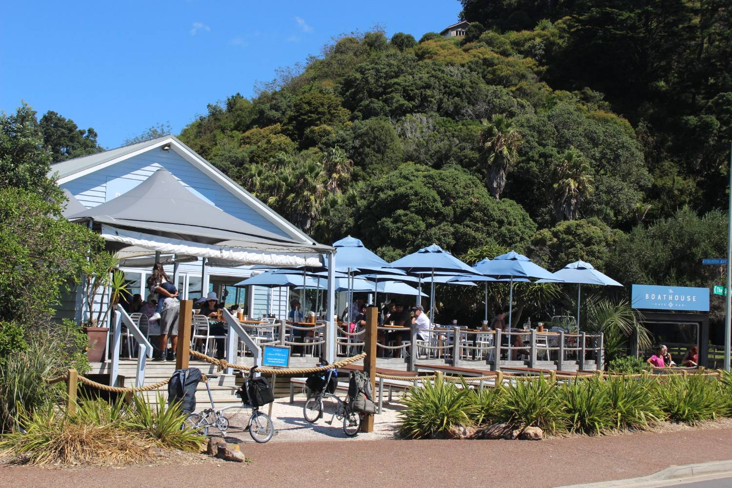Outdoor tables under umbrellas at a café beside the water, bush rising behind