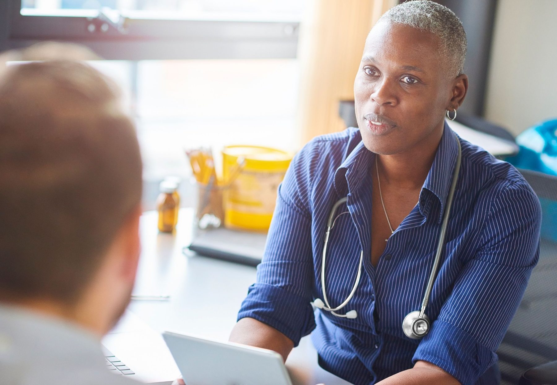 A GP with a stethoscope talking with a patient across her desk