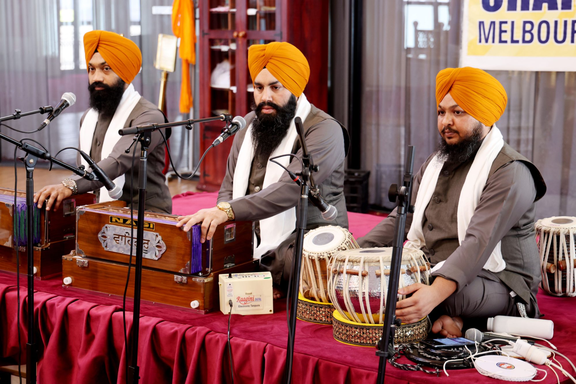 Three musicians in orange turbans performing kirtan at a gurdwara in Melbourne, harmonium and tabla in front of them