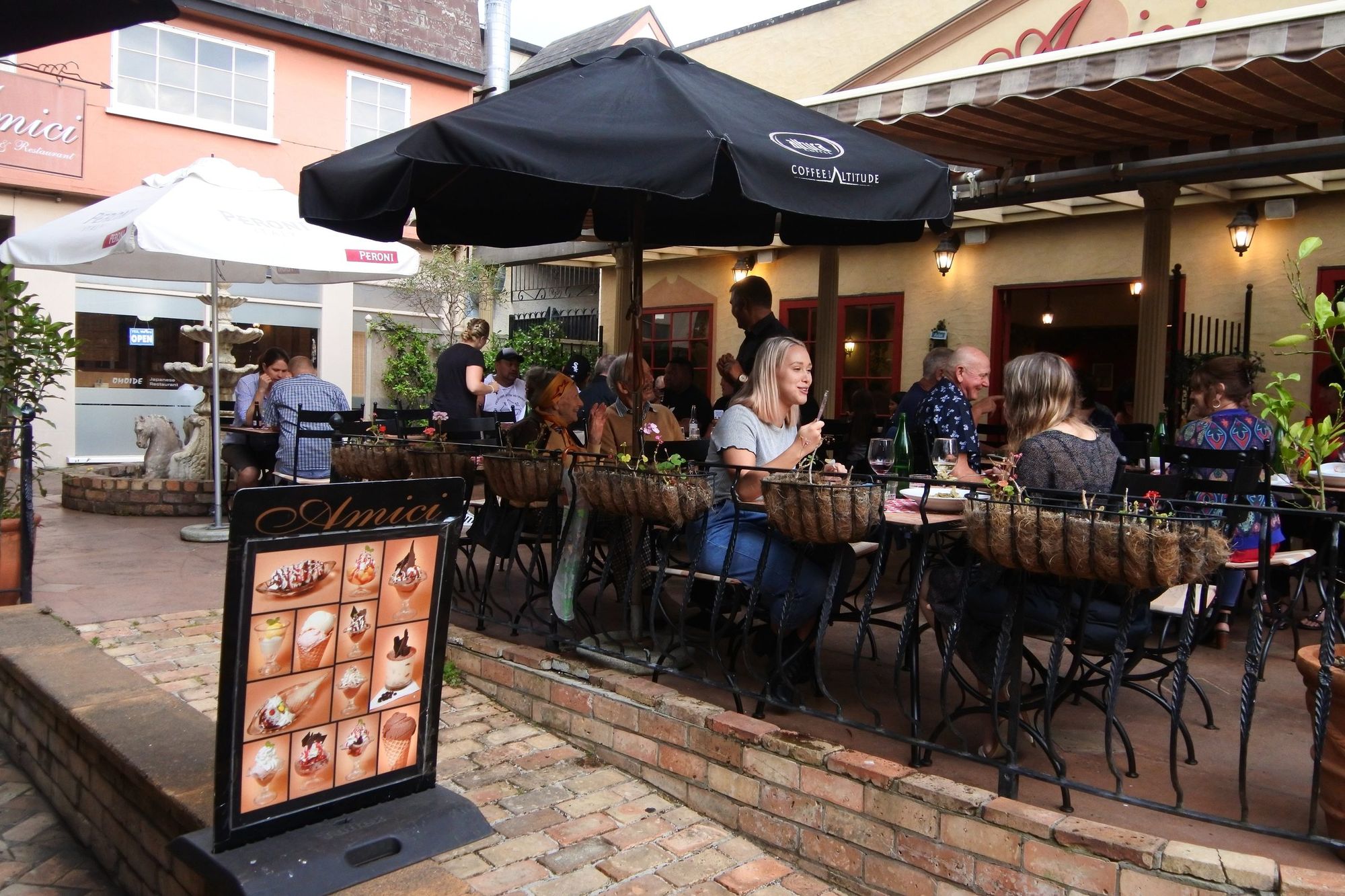 Diners at outdoor tables in a courtyard restaurant in Whāngārei on a warm evening, umbrellas up and every table taken