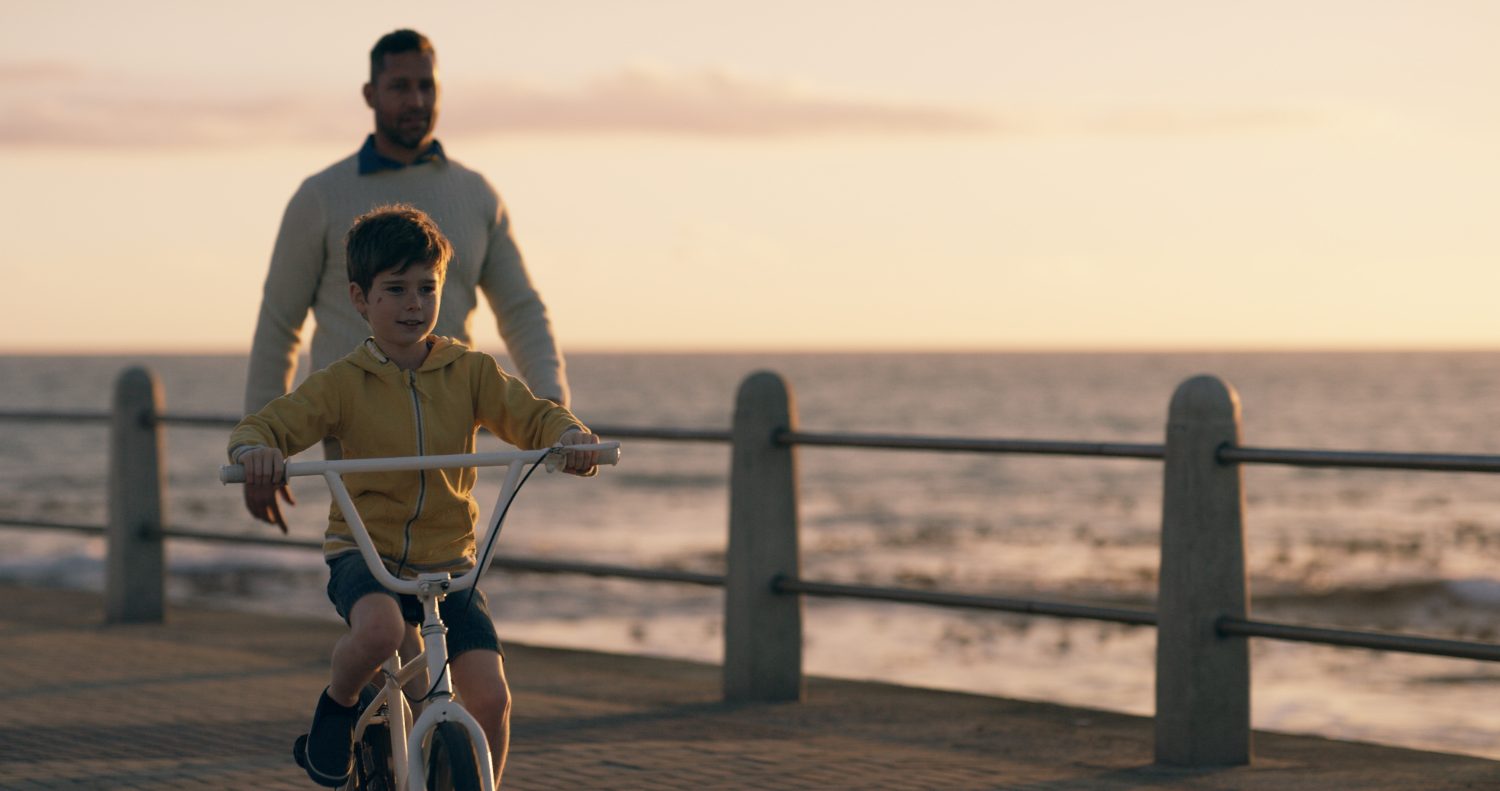 A parent steadying a child on a bicycle beside the water at sunset