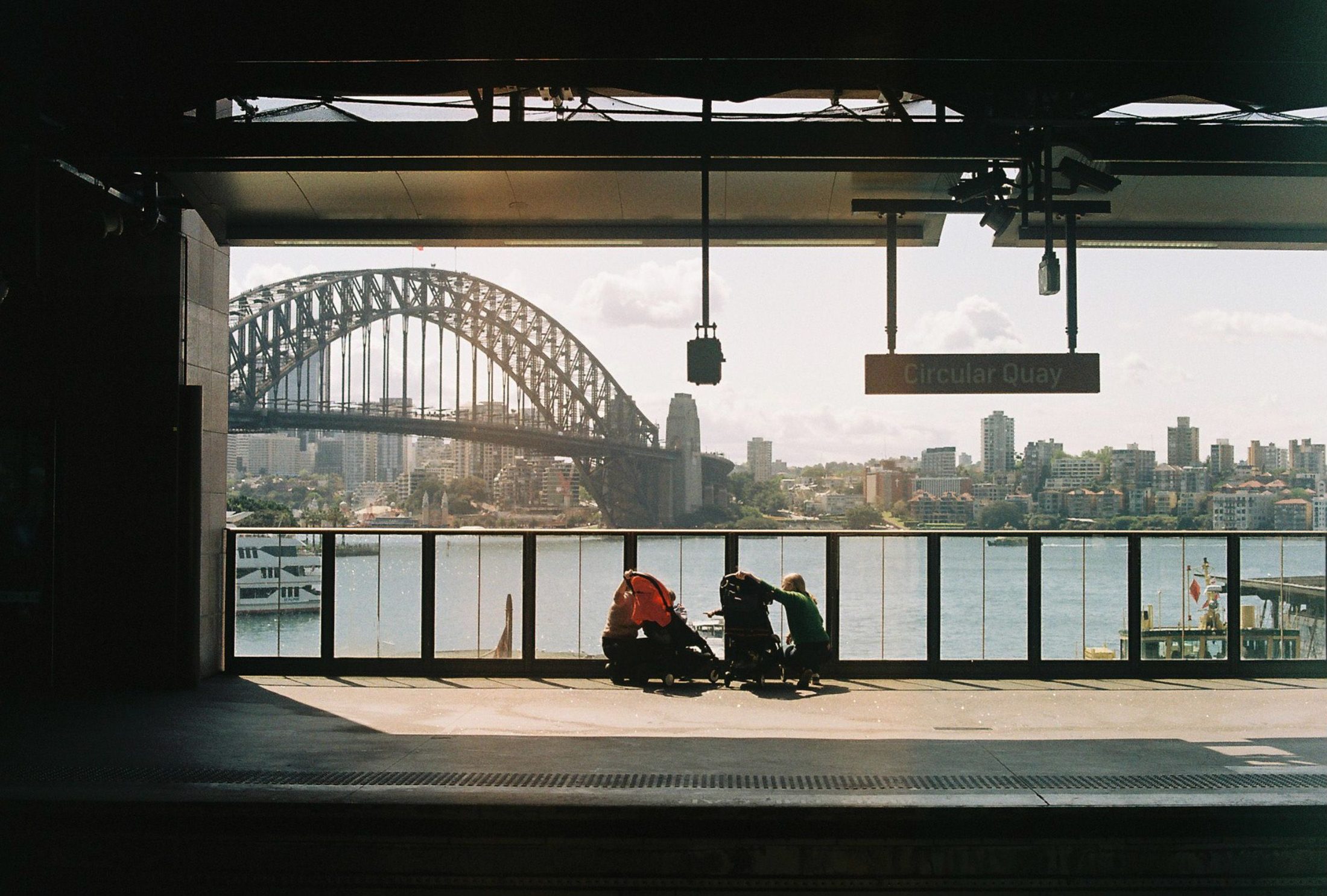 The Harbour Bridge seen through the platform canopy at Circular Quay station, two people with prams waiting at the barrier