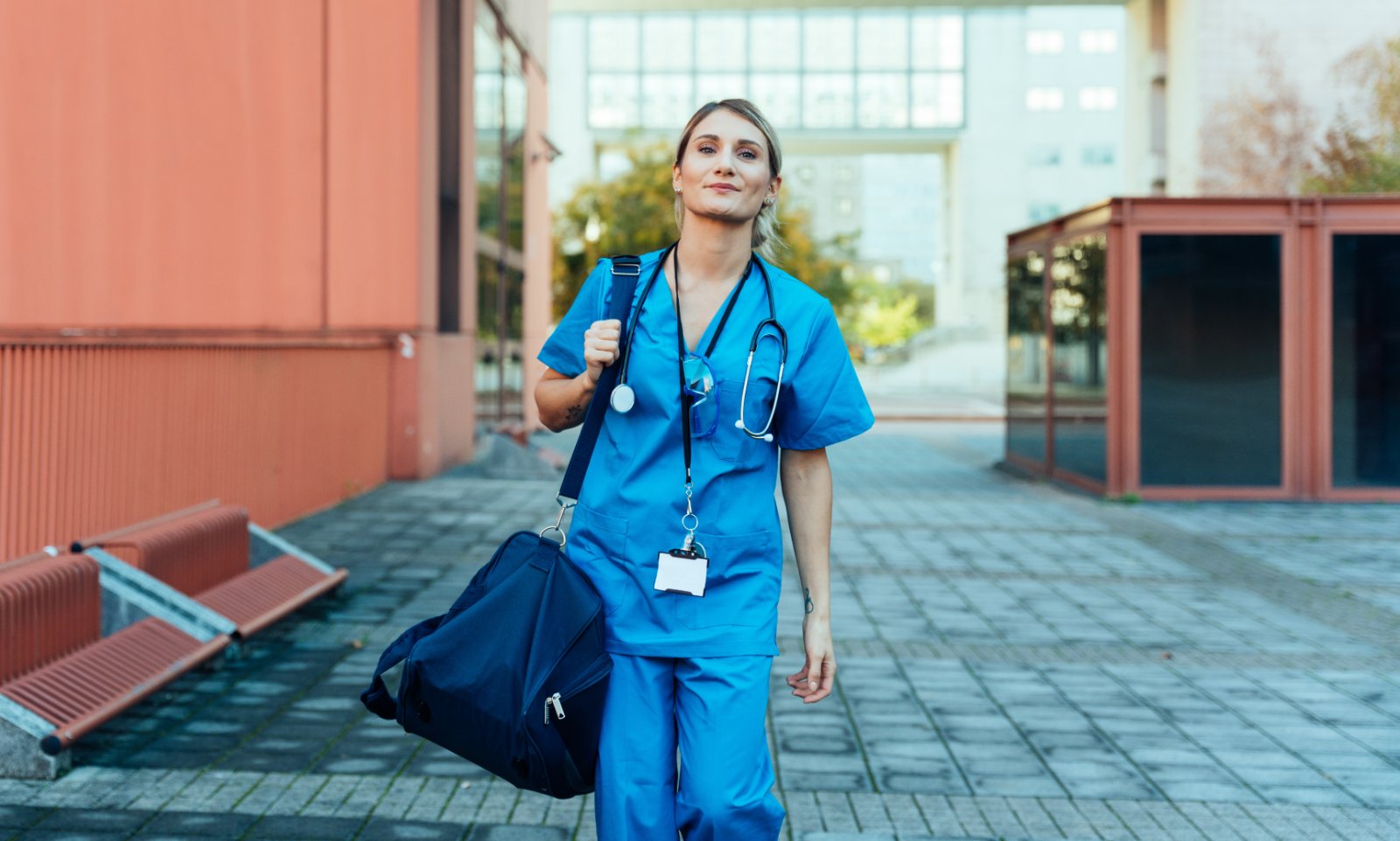 A nurse in scrubs walking up to a New Zealand hospital, bag over her shoulder