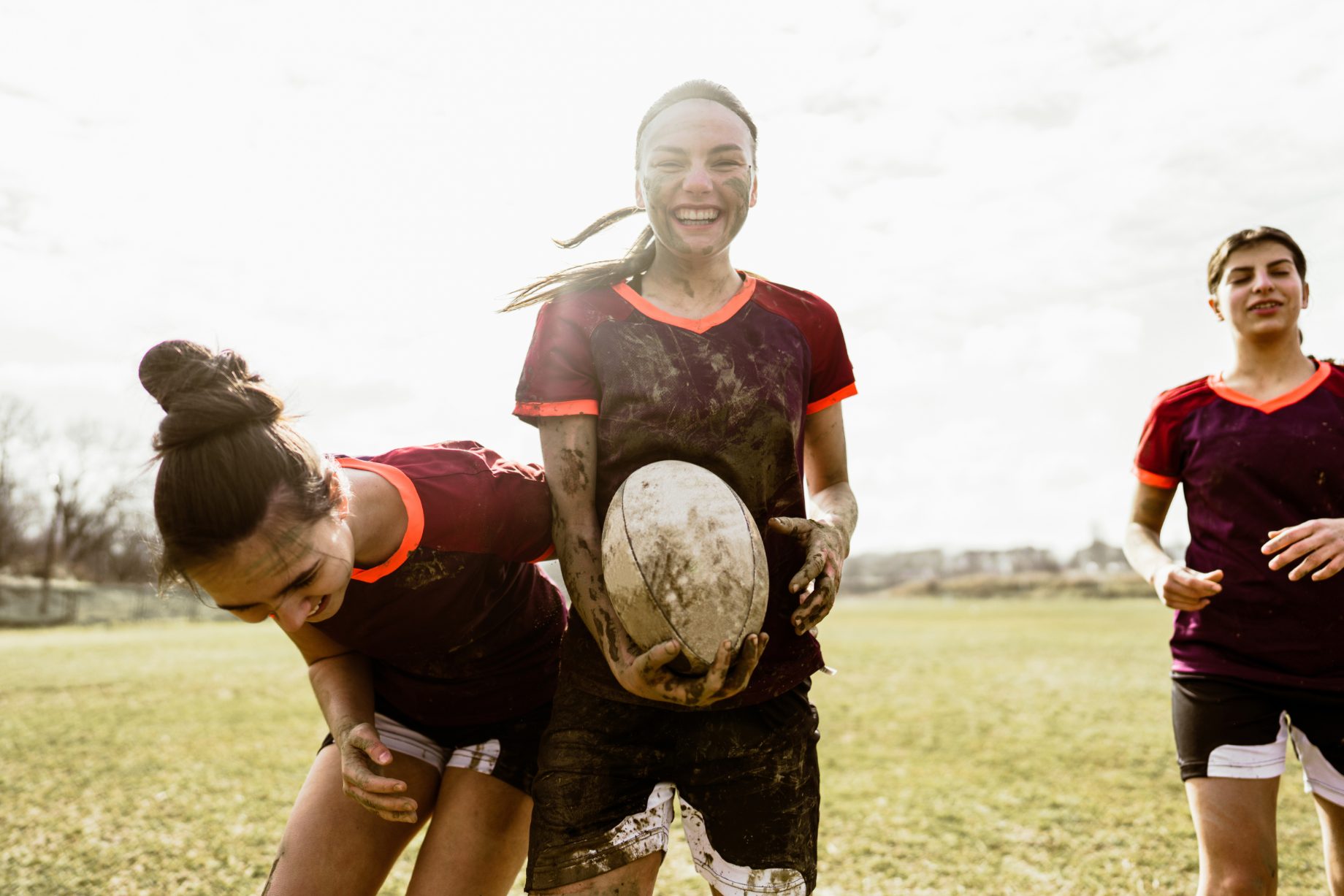 Three rugby players, muddy after a game on a grass pitch, laughing together