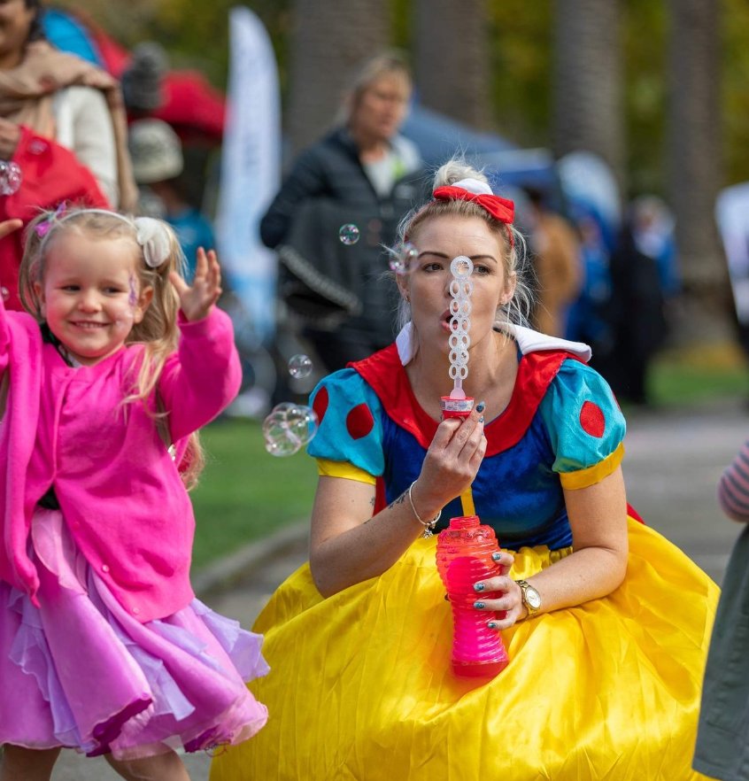 A child in a pink tutu reaching for bubbles blown by a performer in costume at a family festival in Victoria Park, Palmerston North