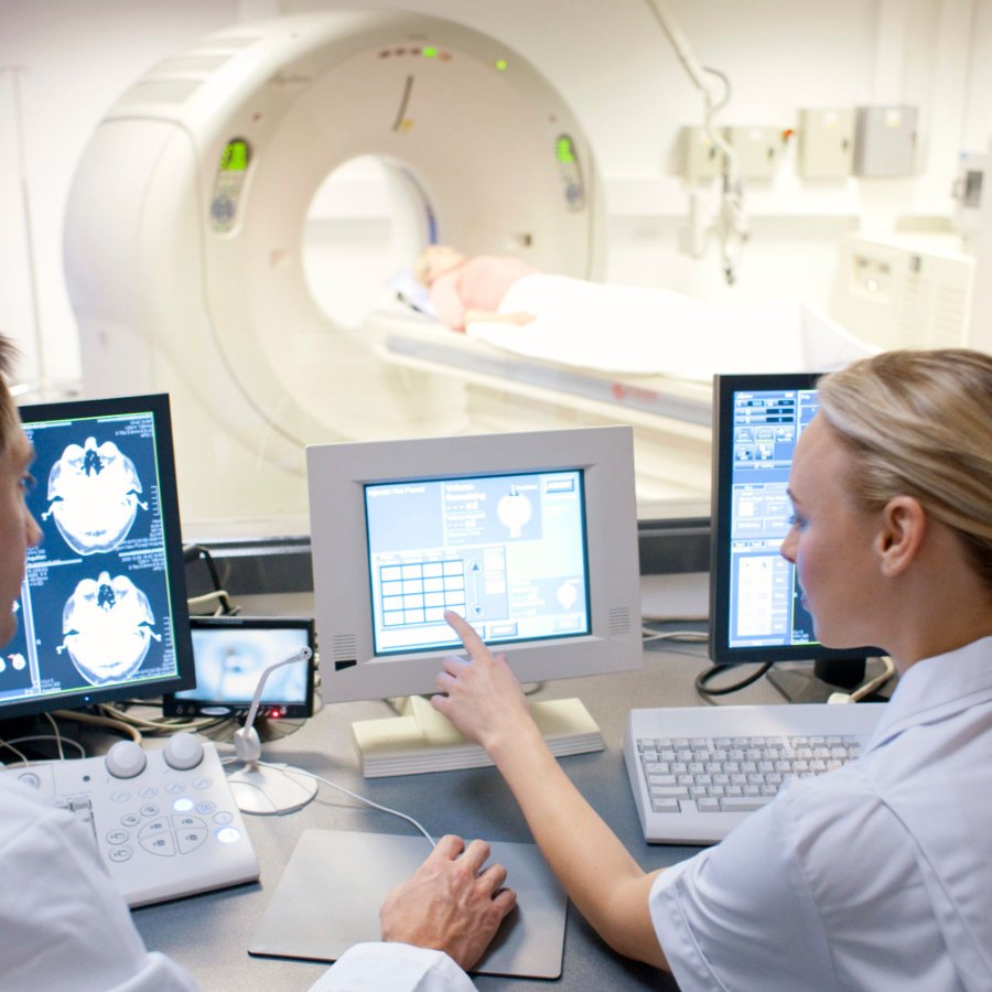 Two clinicians at a control console, scan images on the monitors and the scanner beyond the window