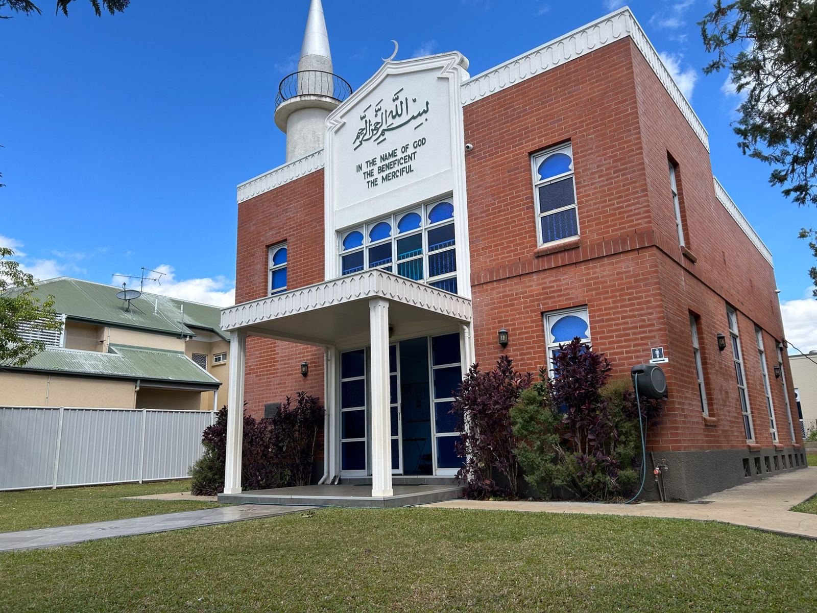 A small-town mosque with a single minaret and blue arched windows, a neighbouring house beside it