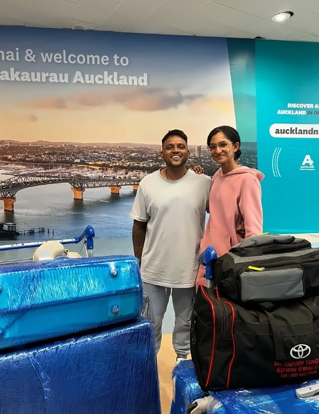 Abigail and her husband standing with their luggage in the Auckland Airport arrivals hall, in front of a mural of the harbour bridge