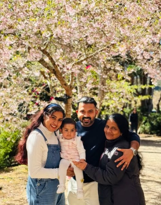 Amal and Sandra holding their baby daughter, smiling with a relative under pink blossom trees