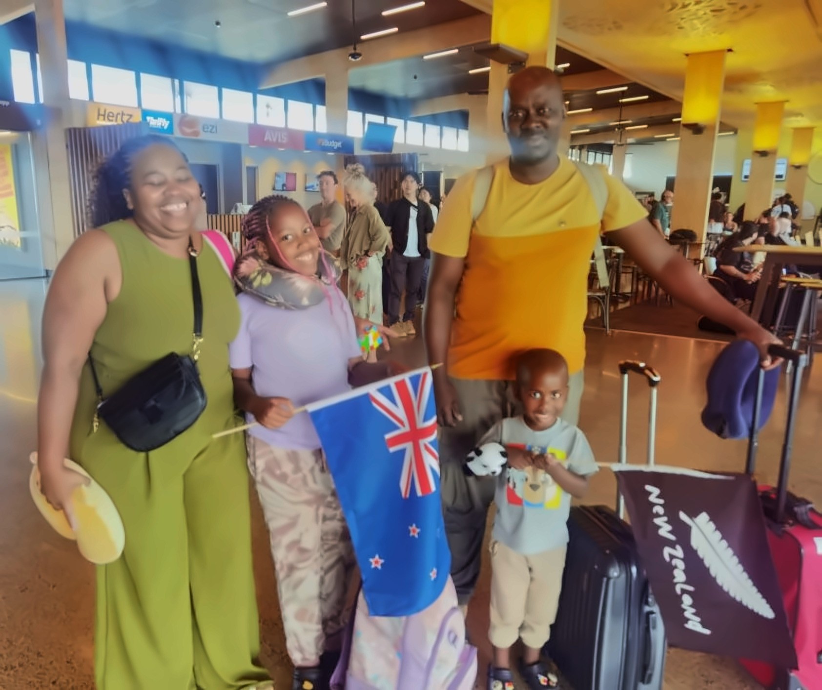 Amanda&rsquo;s husband and two children in the arrivals hall, holding a New Zealand flag and a travel pillow, luggage beside them