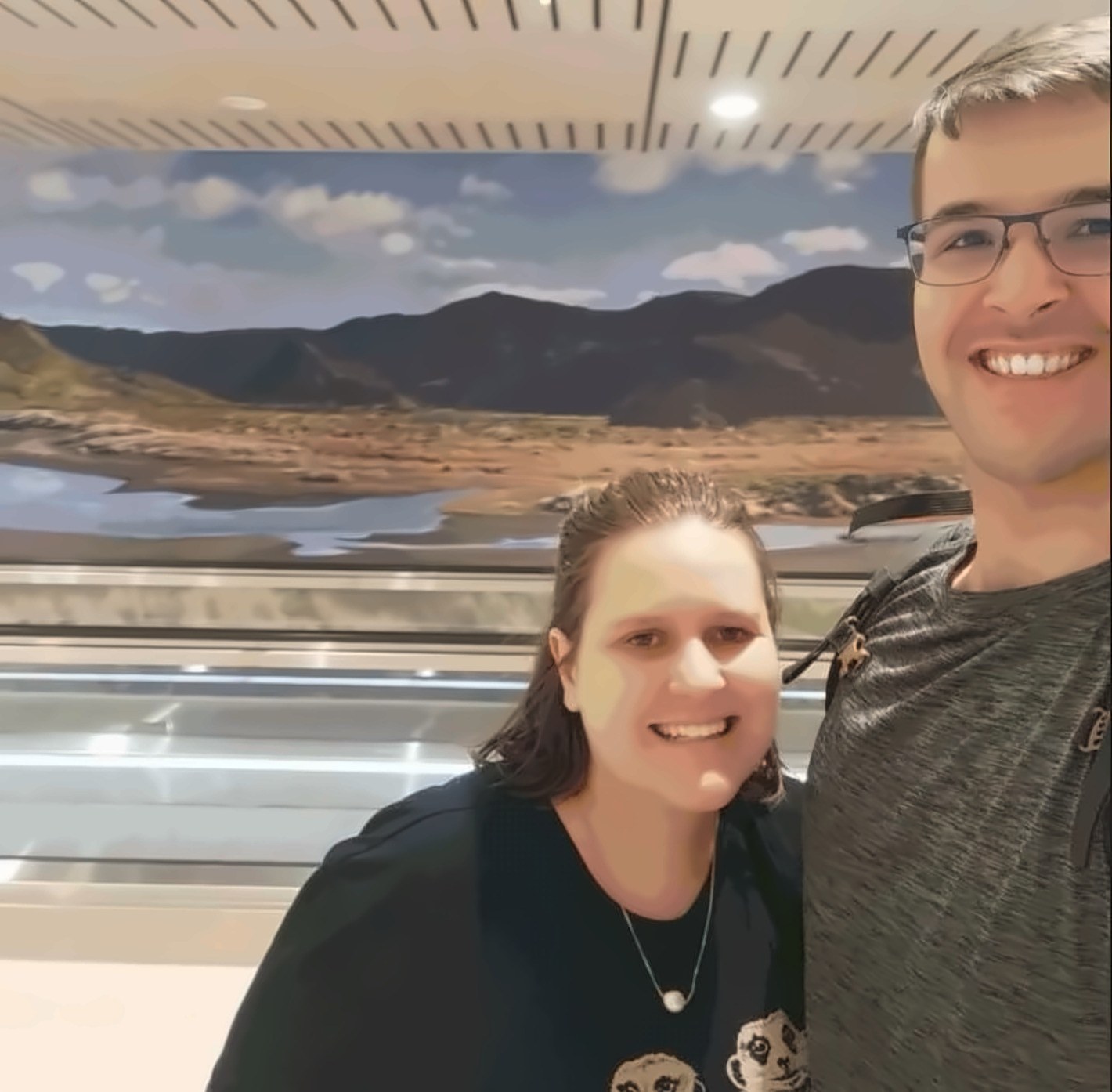 Janita and Jonathan smiling on an escalator, a long photographic mural of a braided river and mountains on the wall behind them