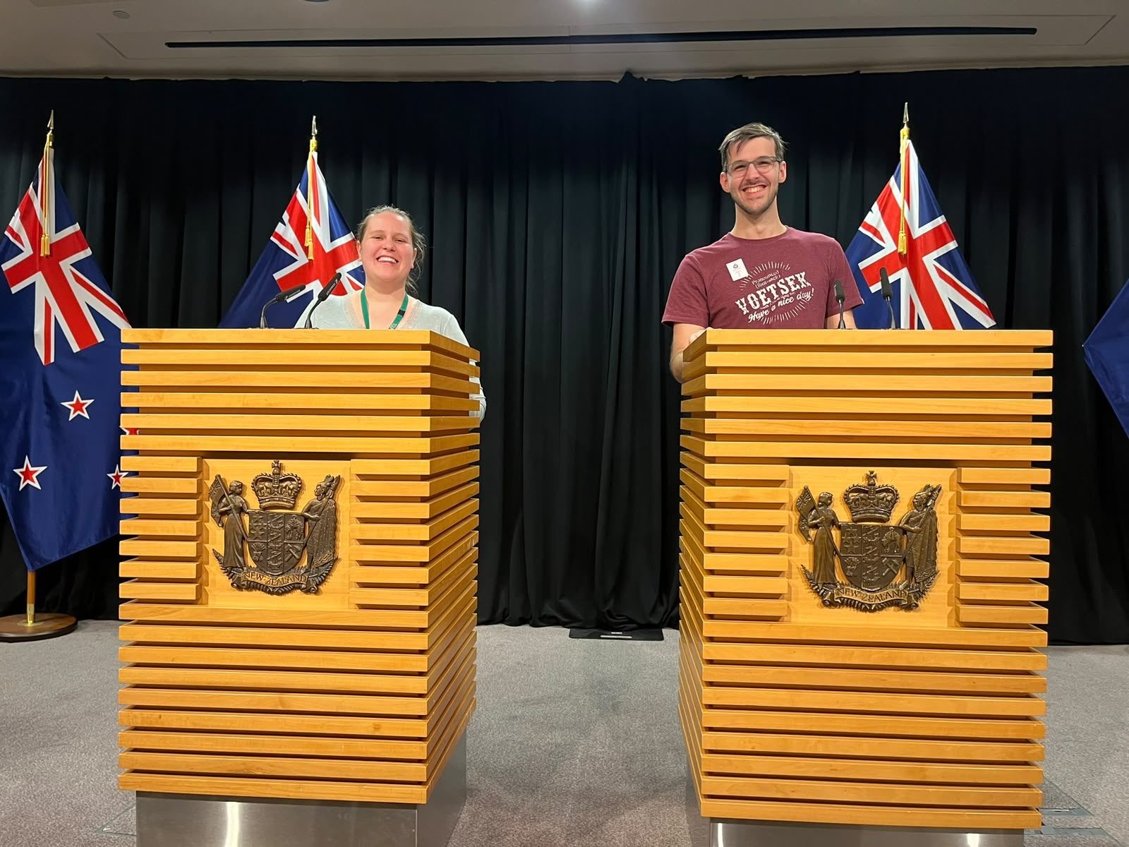 Two visitors behind the wooden lecterns of the Beehive press theatre, New Zealand flags behind them