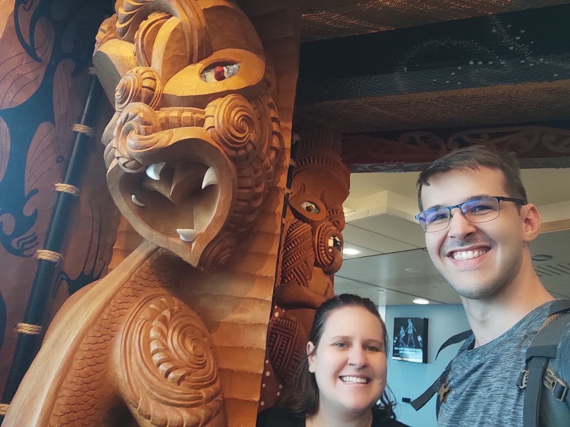 Janita and Jonathan beside a large carved wooden Māori figure with pāua-shell eyes