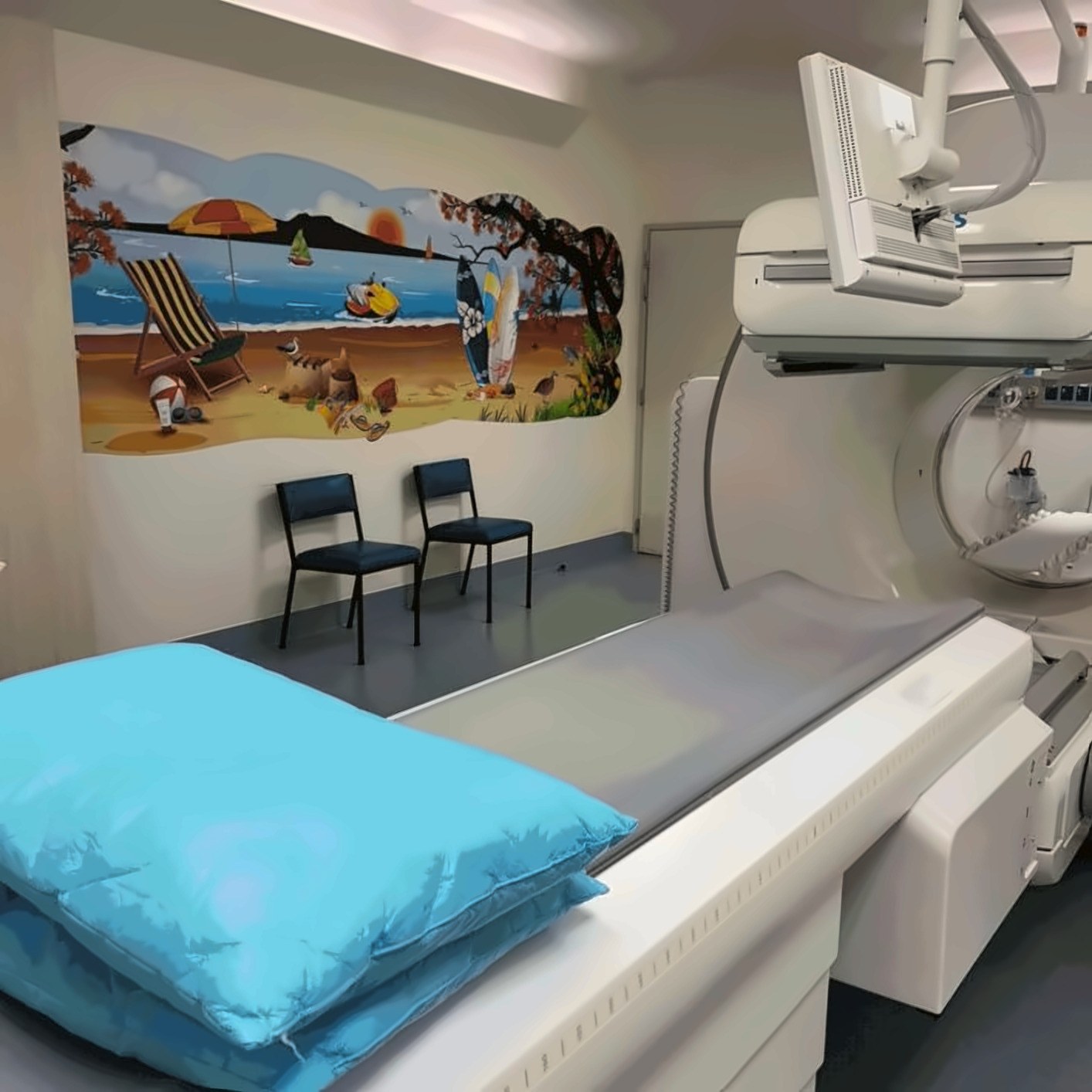 A nuclear medicine scanning room: a gantry and detector head beside a patient couch, with a painted beach mural of deckchairs, surfboards and shorebirds along the wall