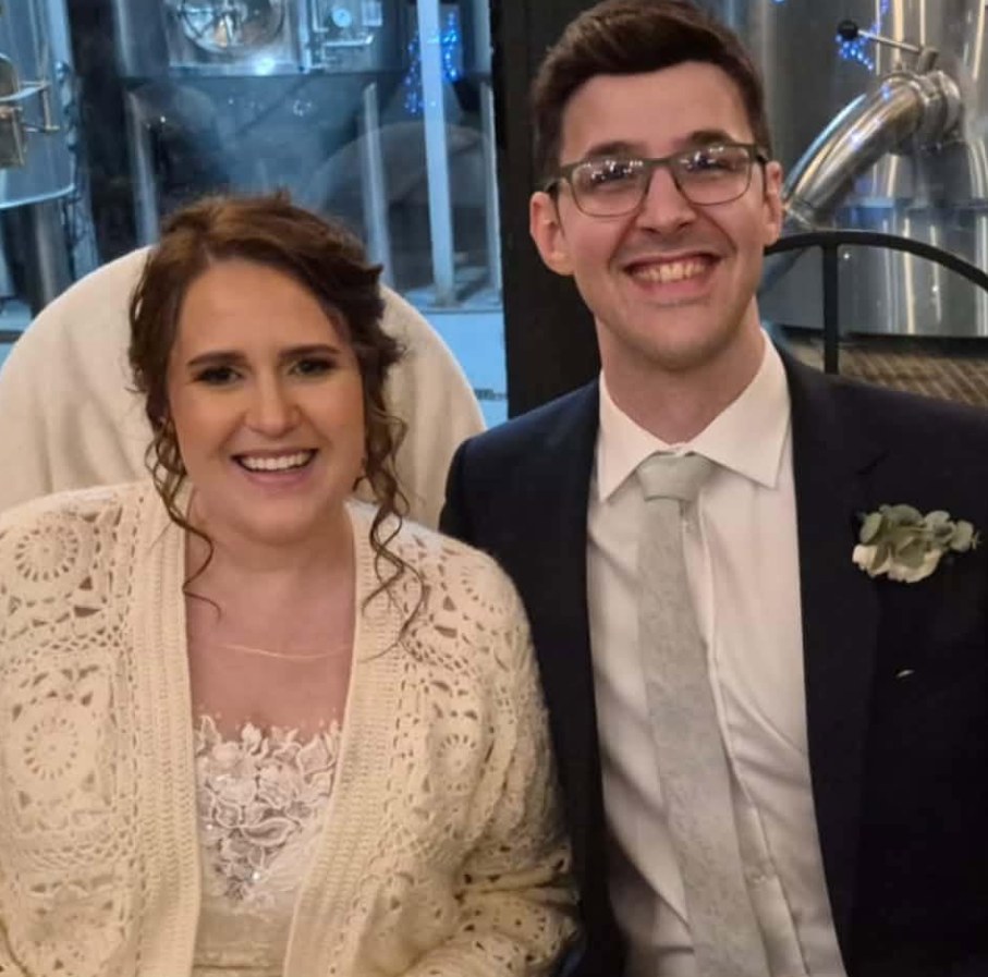 Janita in a lace top and cream crocheted cardigan and Jonathan in a navy suit with a buttonhole, smiling together on their wedding day
