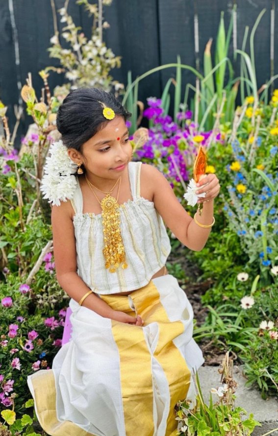 A young girl in a traditional white and gold Kerala outfit with jasmine in her hair, sitting among garden flowers