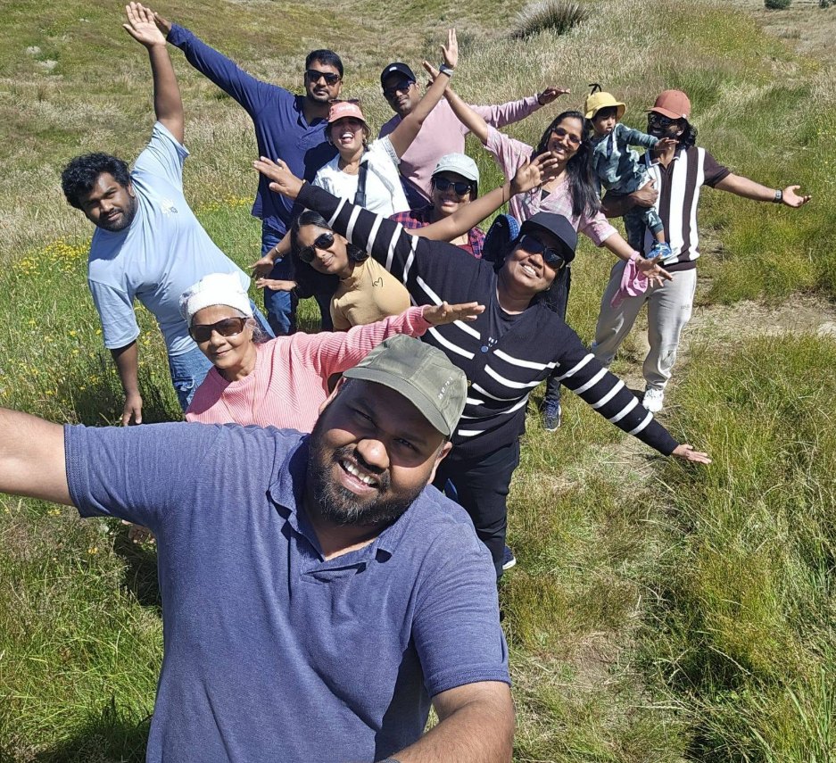 Rezon and friends with their families, arms outstretched for a selfie on a grassy hillside