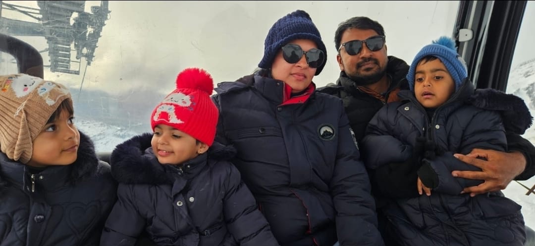 A family with three young children in woolly hats and winter coats riding a gondola above snow