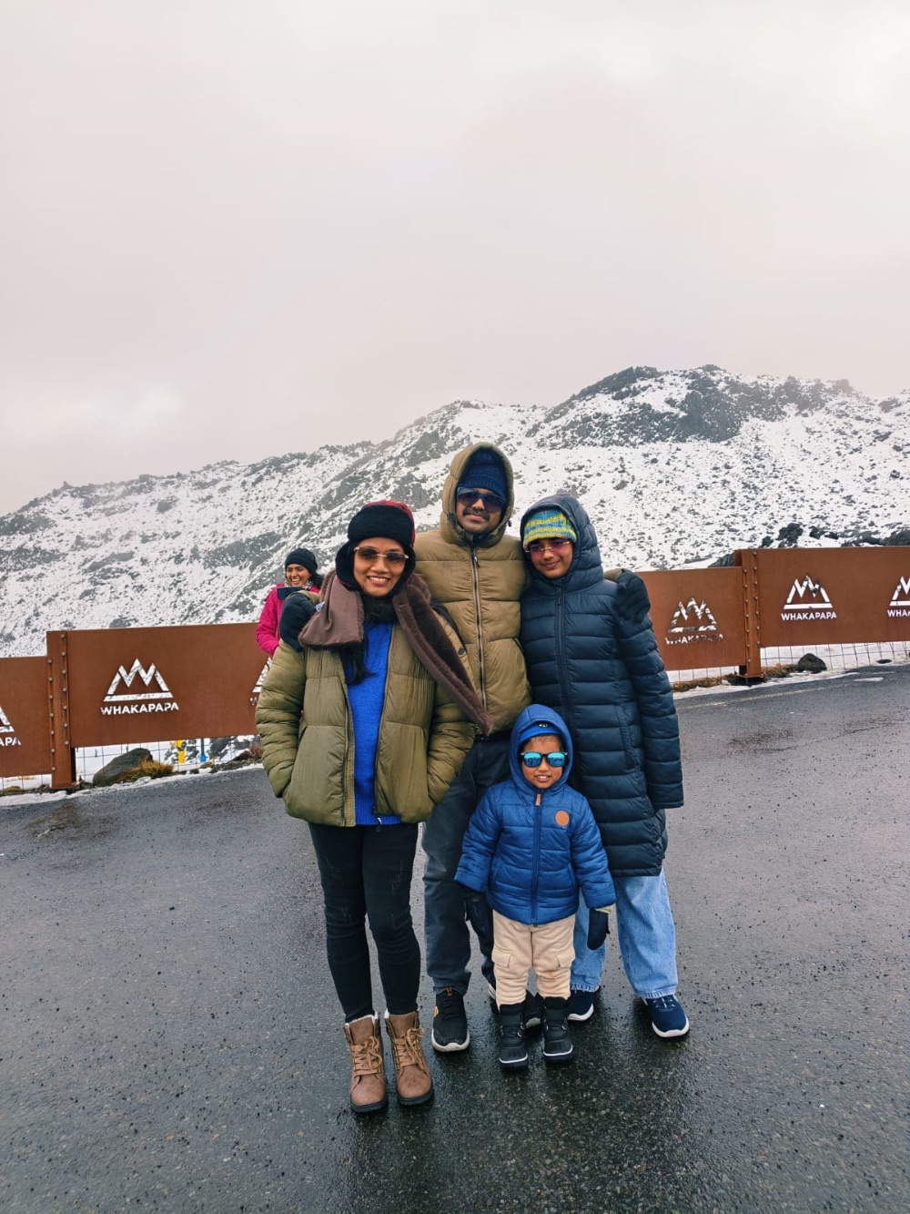 A family in winter coats and hats standing on wet tarmac at a ski area, snow-covered peaks behind them